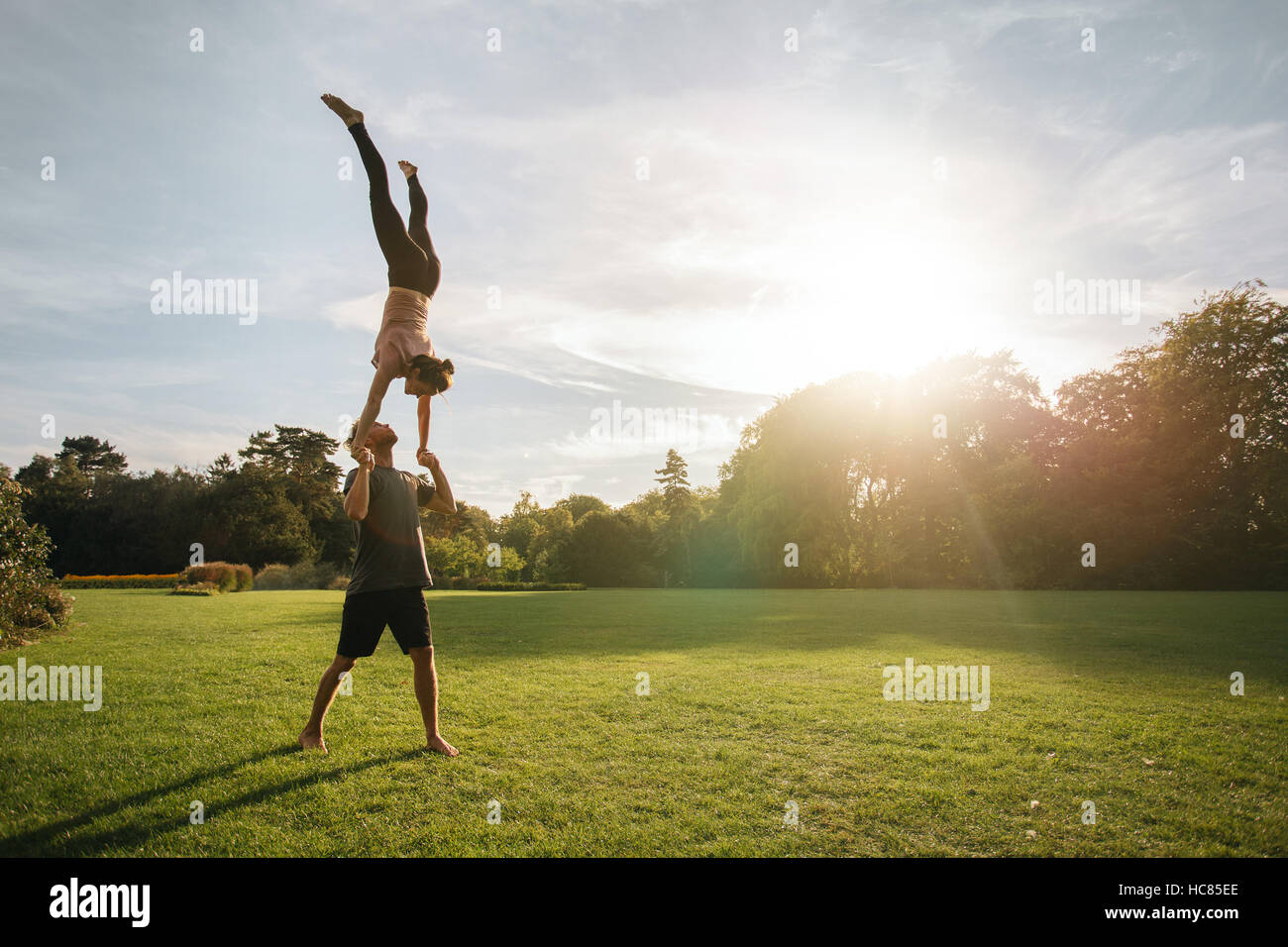 Strong young couple doing pair yoga outdoor in park. Man lifting and balancing woman in park. Acroyoga workout on a sunny morning. Stock Photo