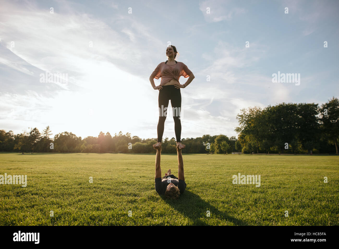 Outdoors shot of young couple doing acrobatic yoga exercise in park. Woman standing on feet of man and balancing. Stock Photo