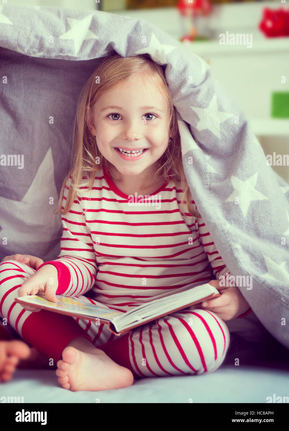 Pretty little girl in sleepwear sitting with book under blanket Stock