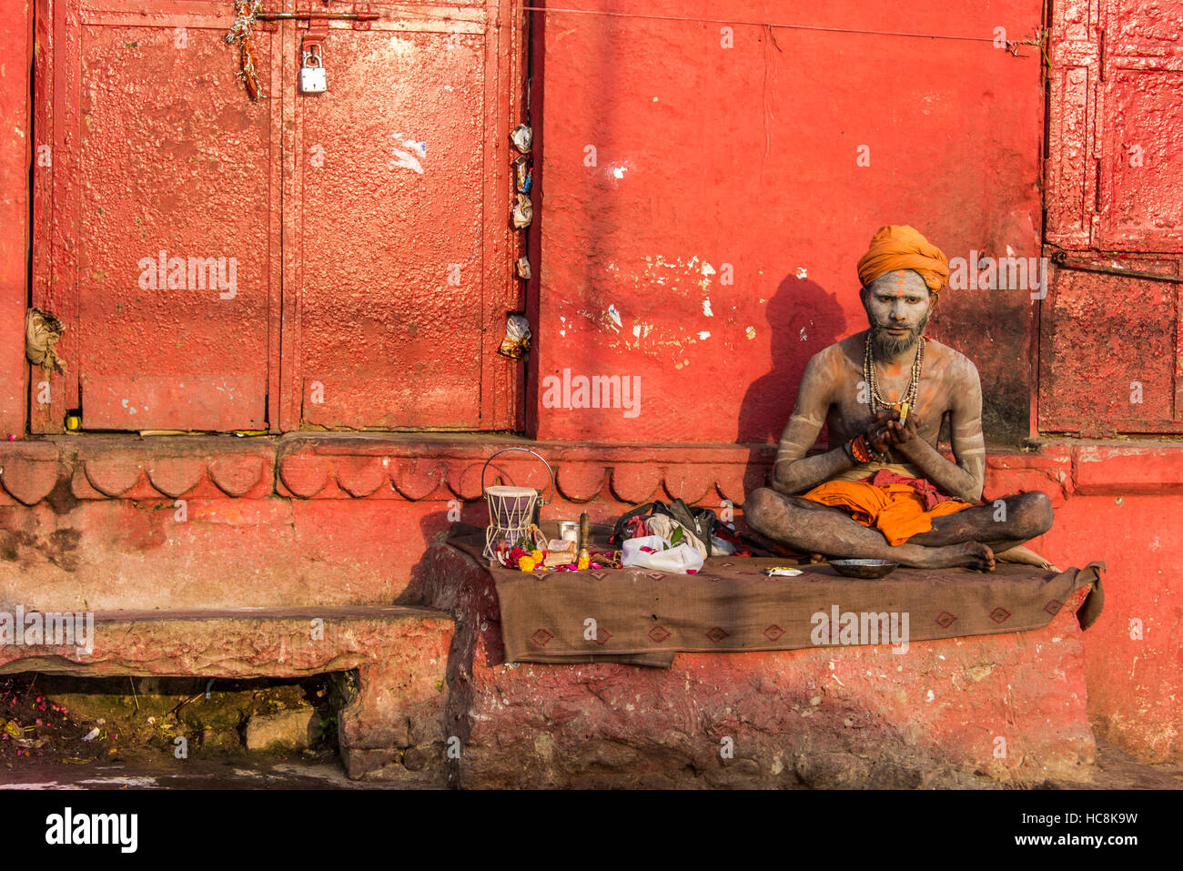 An Indian holy man sits crossed leg on a brown cloth against an orange colored wall with his hands clasped and his head lowered. Stock Photo