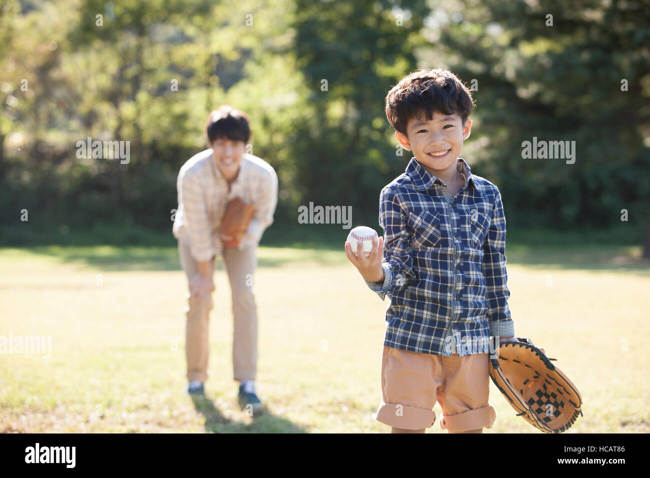 Smiling five-year-old boy with glove and baseball standing on grassland with his father behind him Stock Photo