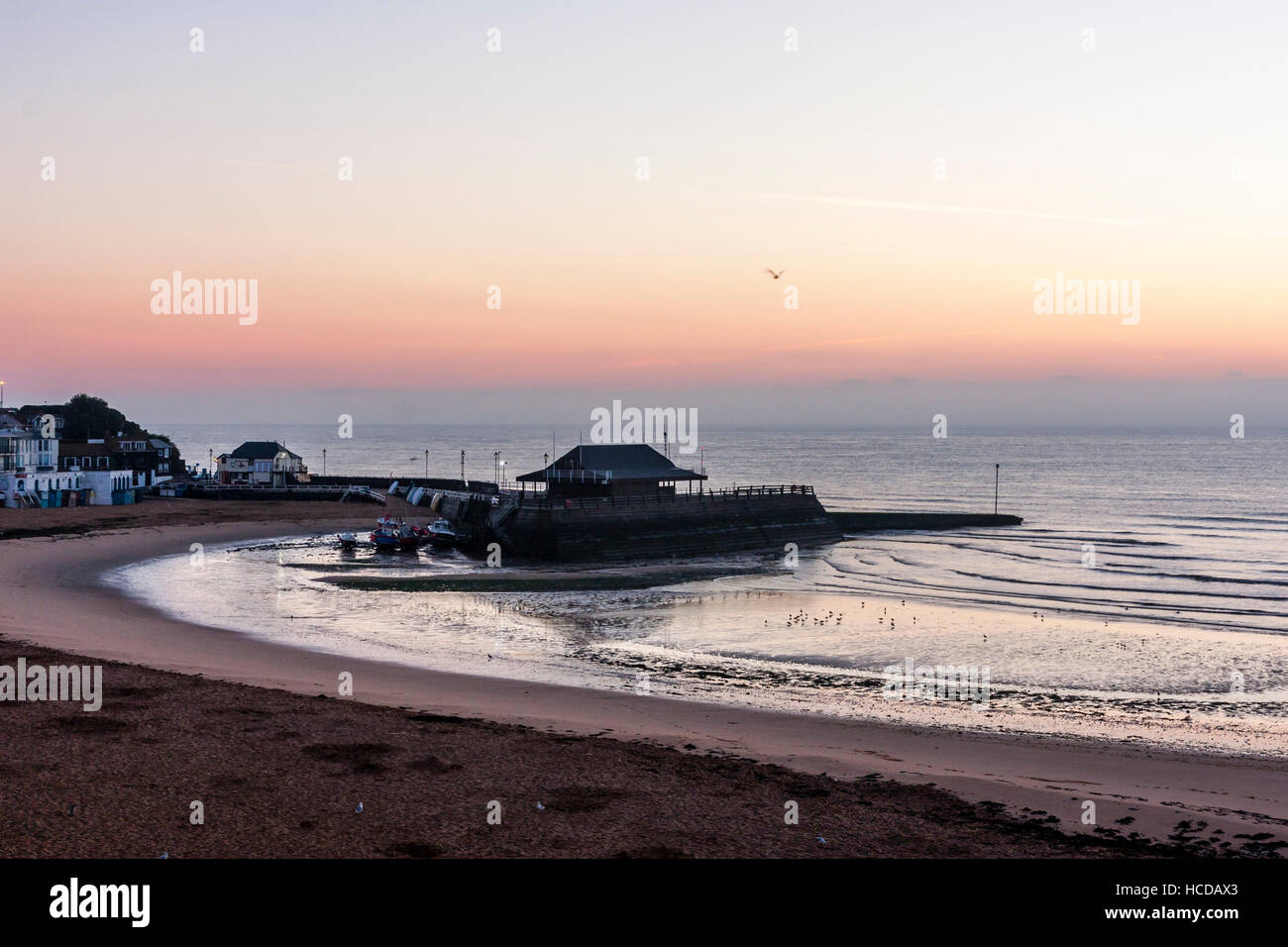 Dawn sky breaking over the sea at Broadstairs with the harbour and beach in the foreground. Clear sky with cloud layer on horizon. Calm sea. Stock Photo
