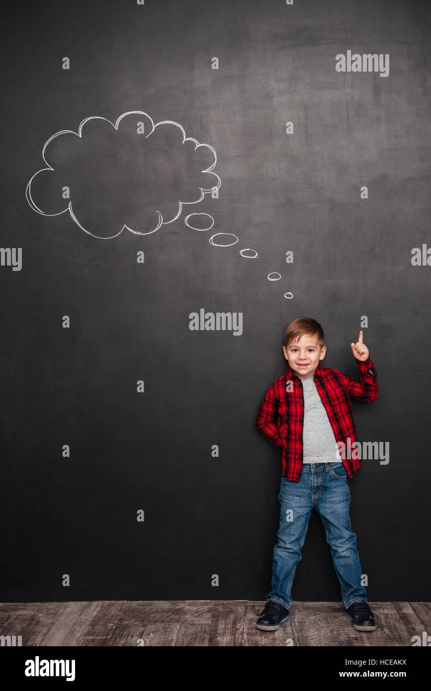 Child thinking with a thought bubble on the chalkboard concept for confusion, inspiration and solution while pointing up. Stock Photo