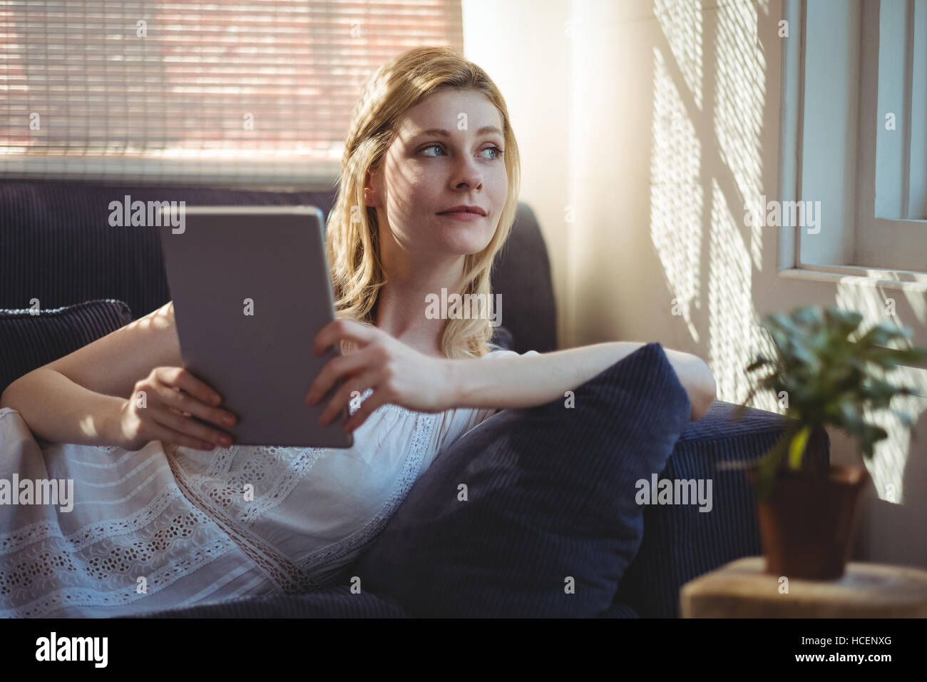 Thoughtful woman lying on sofa and holding digital table in living room Stock Photo