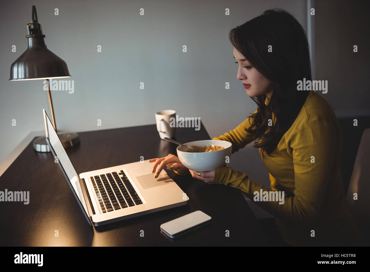 Woman eating cereal while working on laptop in study room Stock Photo