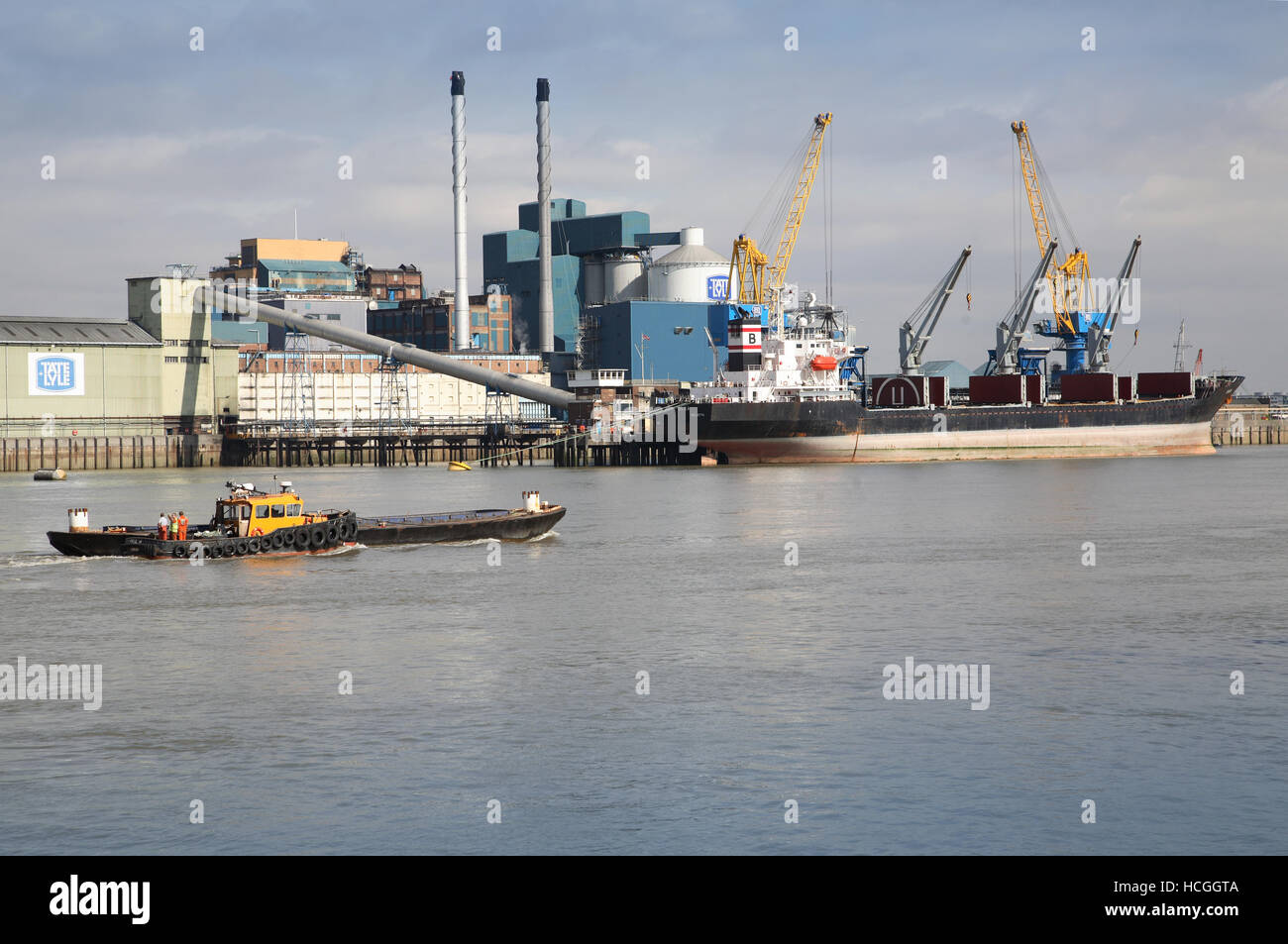 A barge passes the Tate and Lyle sugar refinery next to the River Thames at Silvertown in London's Docklands. Shows ship moored. Stock Photo