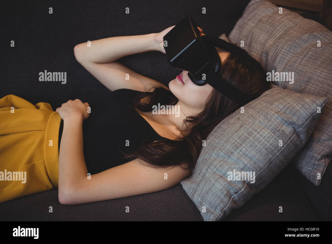 Woman using virtual reality headset while lying on sofa in living room Stock Photo