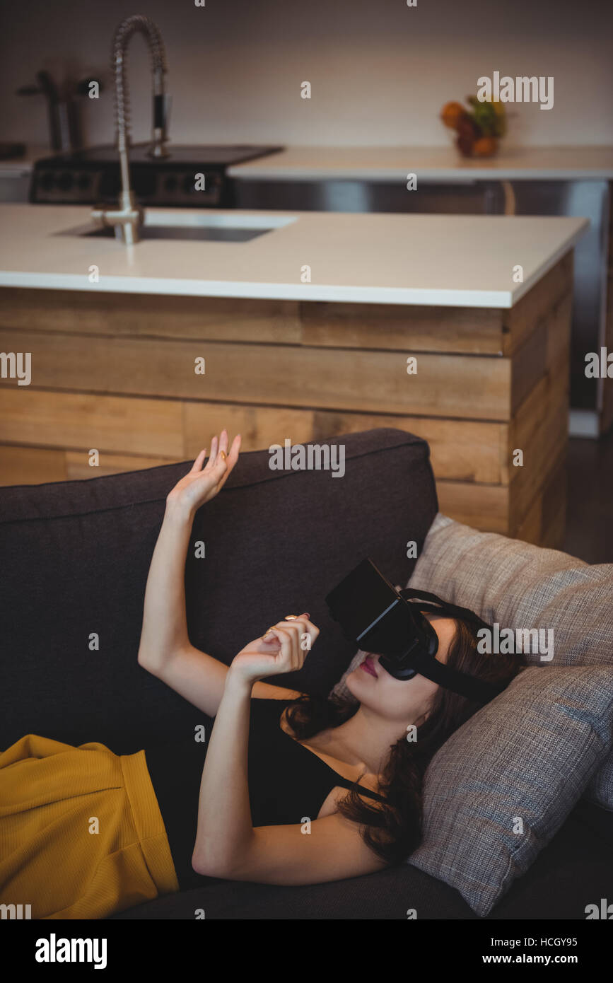 Woman using virtual reality headset while lying on sofa in living room Stock Photo