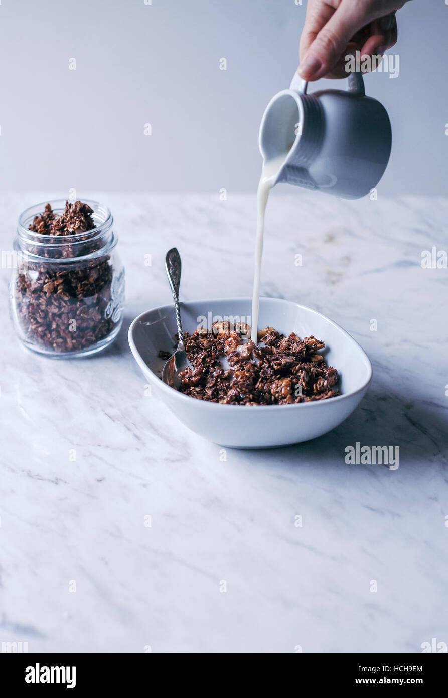 Woman pouring almond milk into a bowl of homemade chocolate granola with oats, nuts and seeds Stock Photo