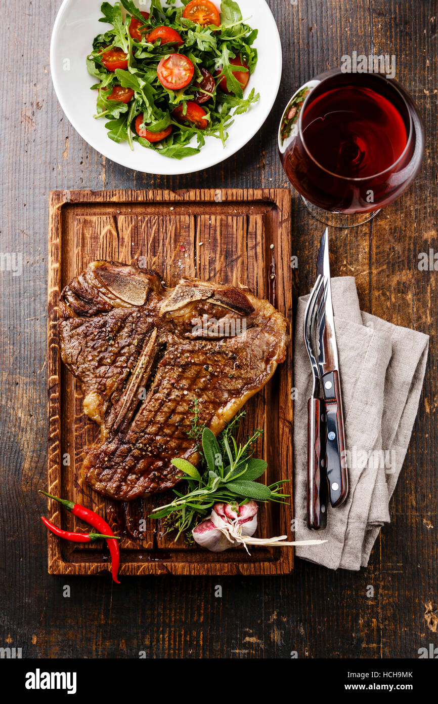 Grilled T-Bone Steak with Salad with Cherry tomatoes and Arugula on serving board on wooden background and red wine Stock Photo