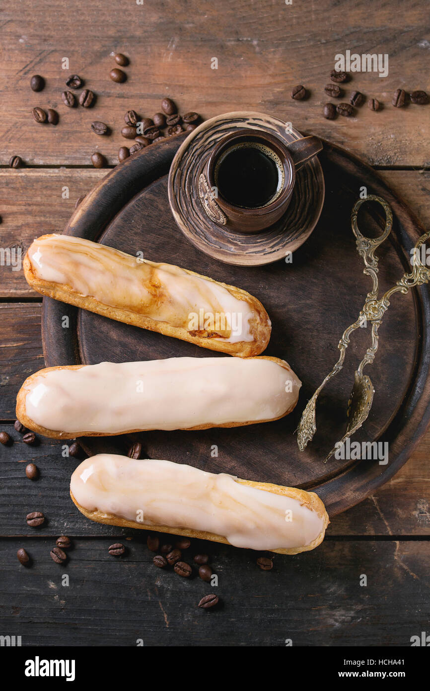 Fresh homemade eclairs with different glaze, coffee beans and ceramic cup of black coffee over old wooden texture background with space for text. Top Stock Photo
