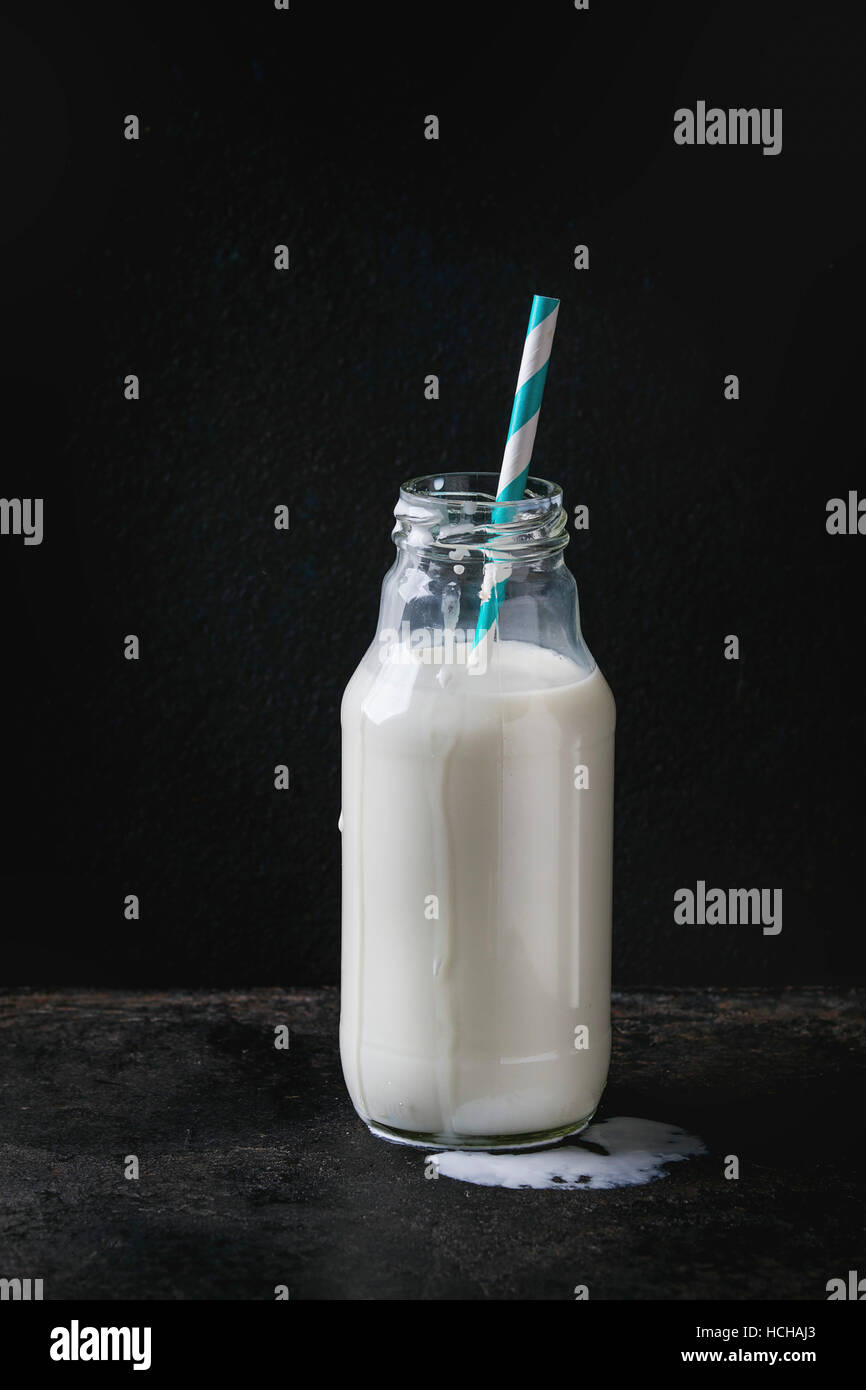 Opened glass bottle full of milk with spot of milk and striped retro cocktail tube over black background. Copy space Stock Photo