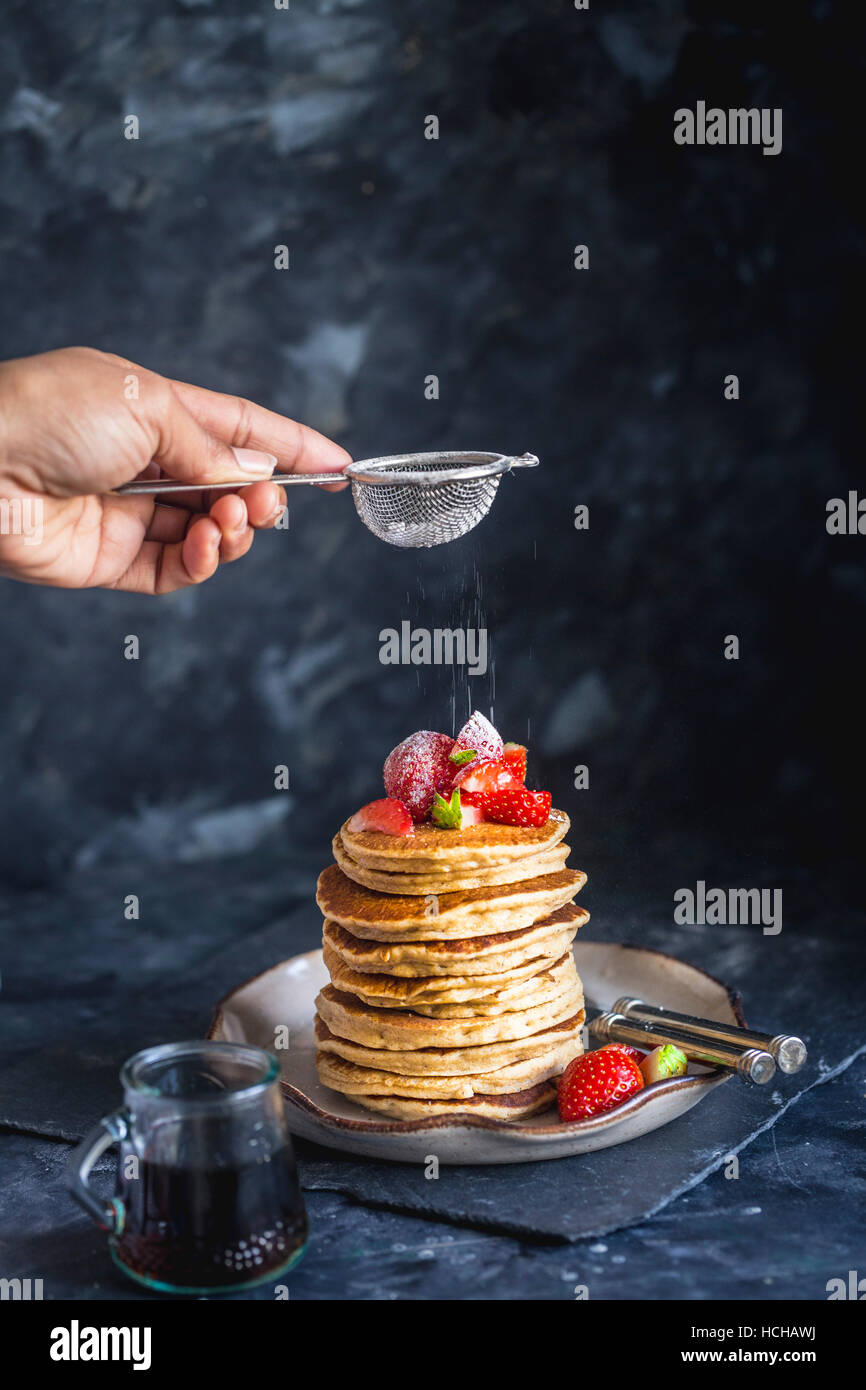 Dusting Icing Sugar on Stack of Gluten free Oat Flour Pancakes served with chopped Strawberries and Maple Syrup Stock Photo