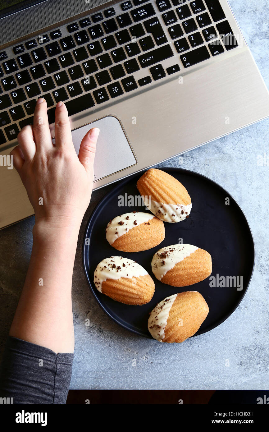 Female sitting at a table,working on the laptop computer and eating madeleines cookies.Top view Stock Photo