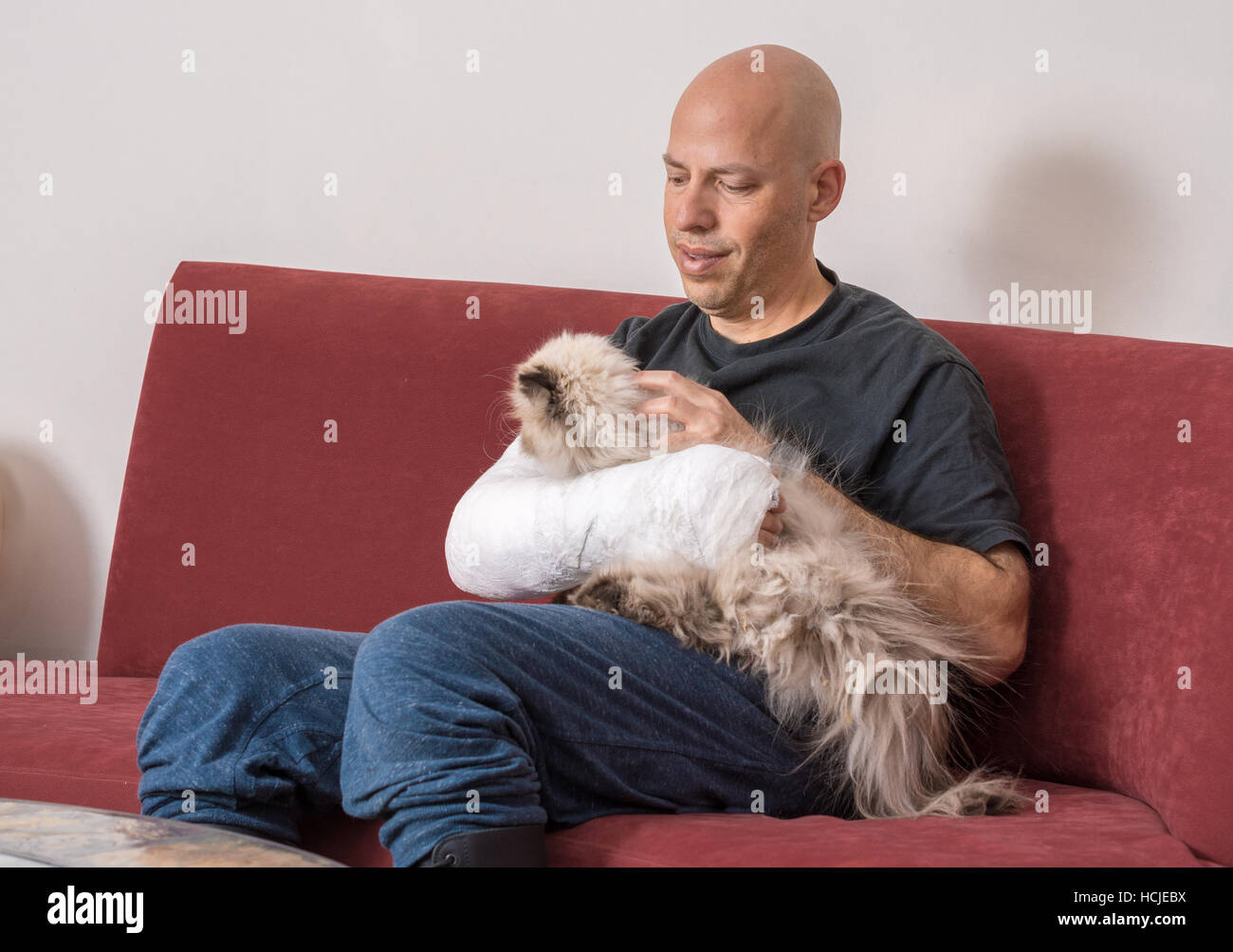 Young man with an arm and elbow in a white plaster / fiberglass cast at home, relaxing while  petting his cat  after having broken his  arm Stock Photo