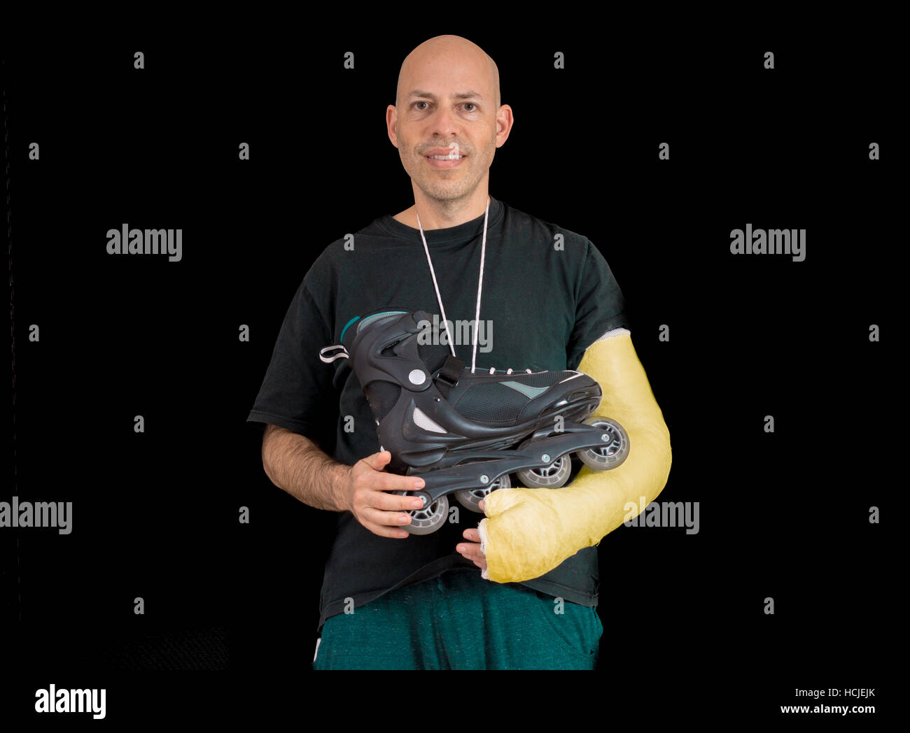 Young man sporting a bright yellow arm cast, holding an inline skate after a breaking his arm in a rollerblading accident, isolated on black Stock Photo