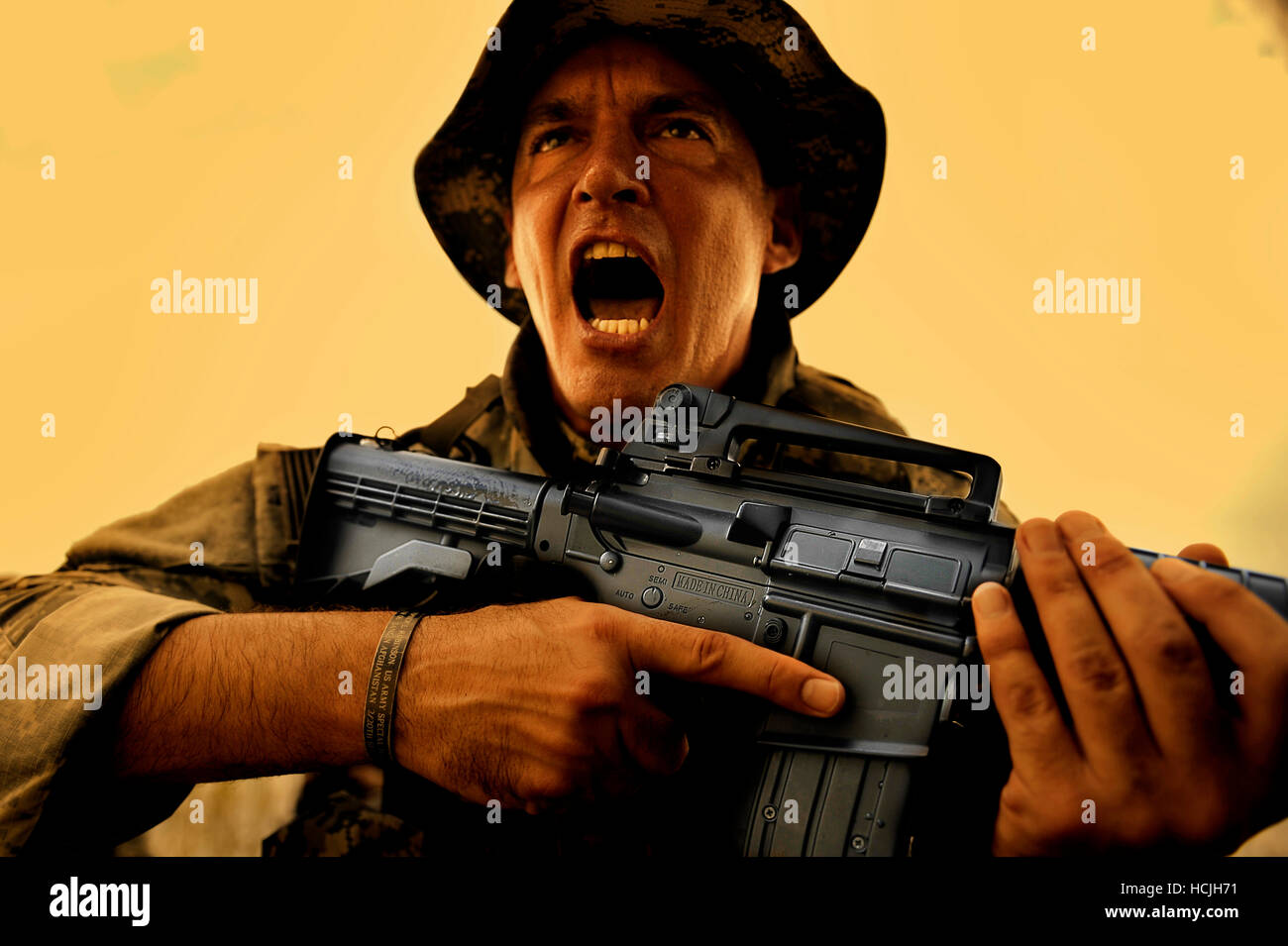 A soldier screams loudly as he demonstrates how to use a machine gun. Wearing a boonie cap and military uniform with the sleeves rolled up, he clutches an automatic rifle. Stock Photo