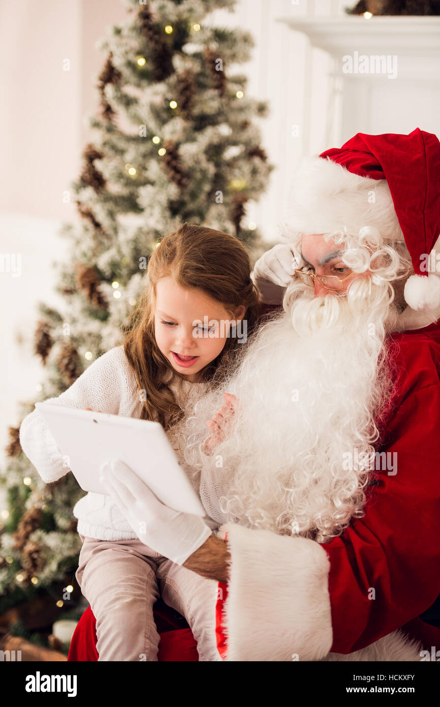 Little girl sitting with Santa using tablet on the armchair at home ...