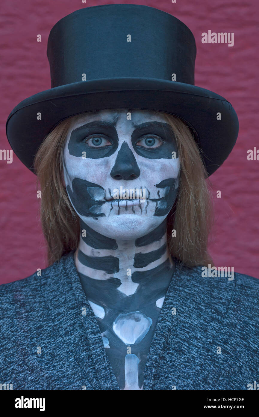 Girl wearing top hat, face painted as death, in front of red wall