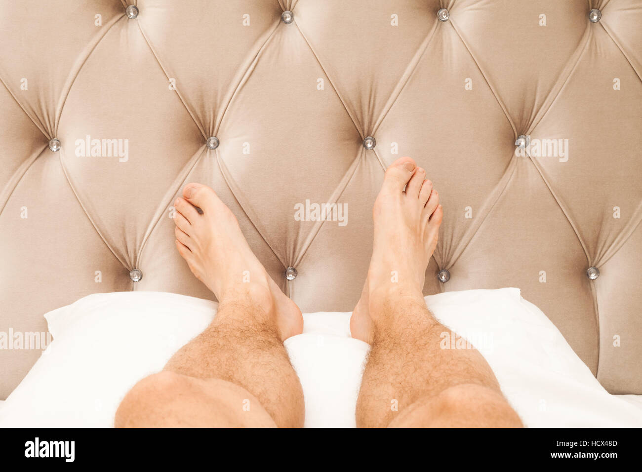Relaxing male feet lay on white pillow near soft headboard in bedroom, luxury rest lifestyle Stock Photo