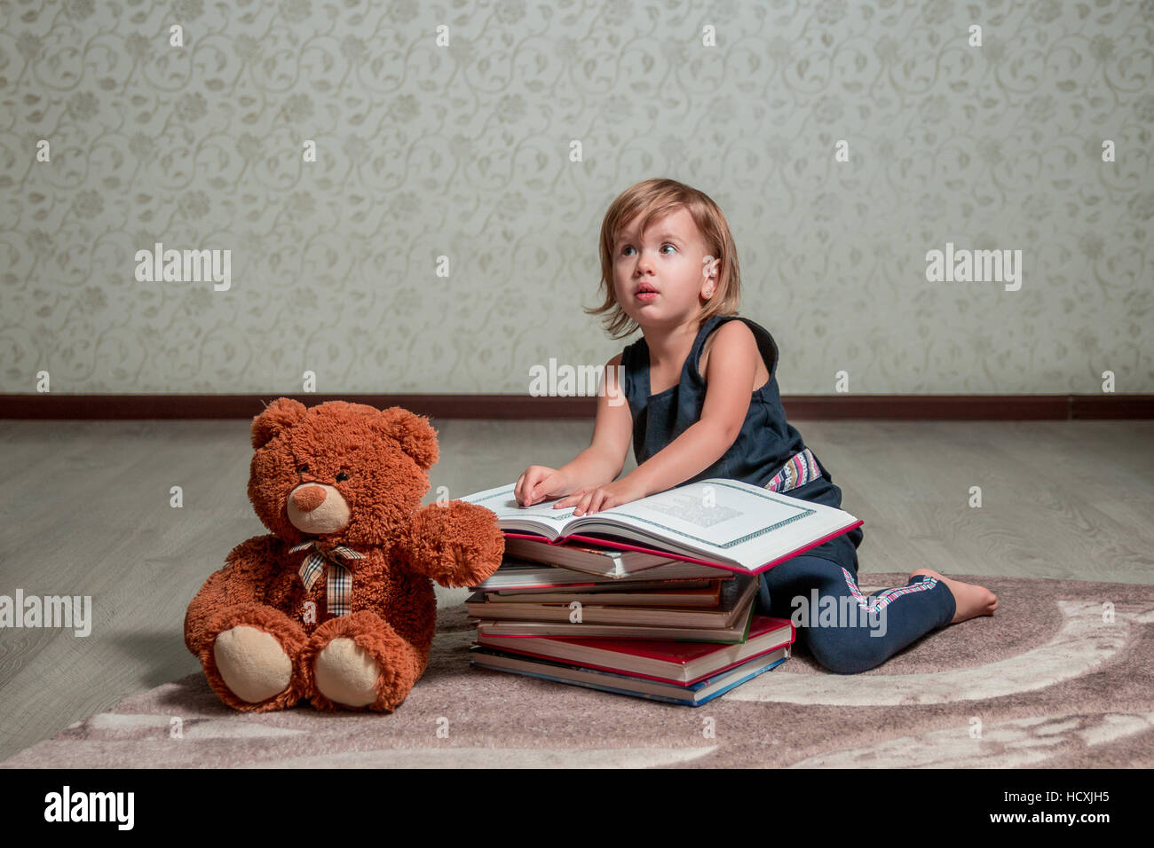 A little girl in a dark blue dress reading a book sitting on the floor ...