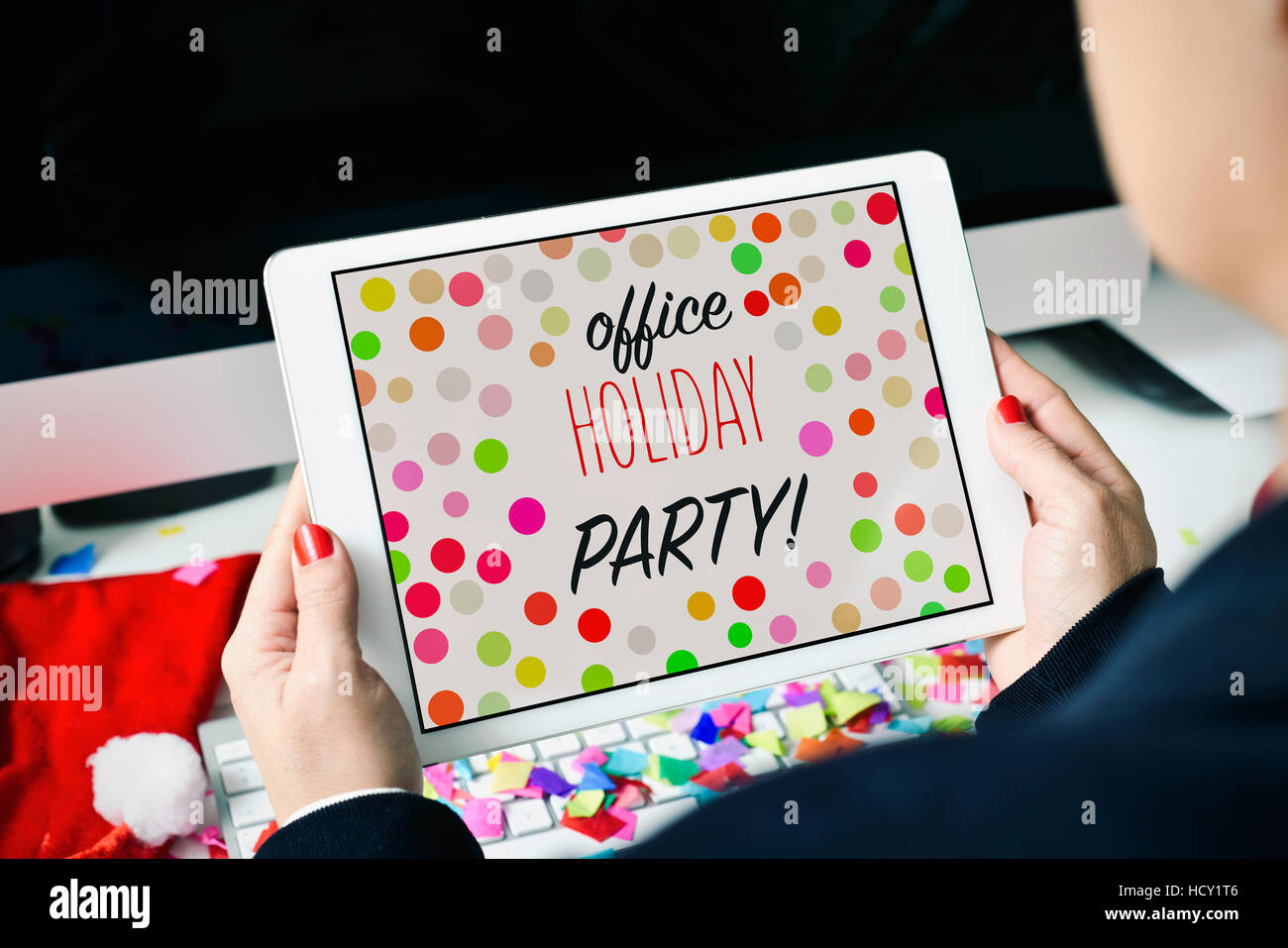 closeup of a young caucasian woman sitting at her office desk full of confetti looking to her tablet, with the text office holiday party in its screen Stock Photo