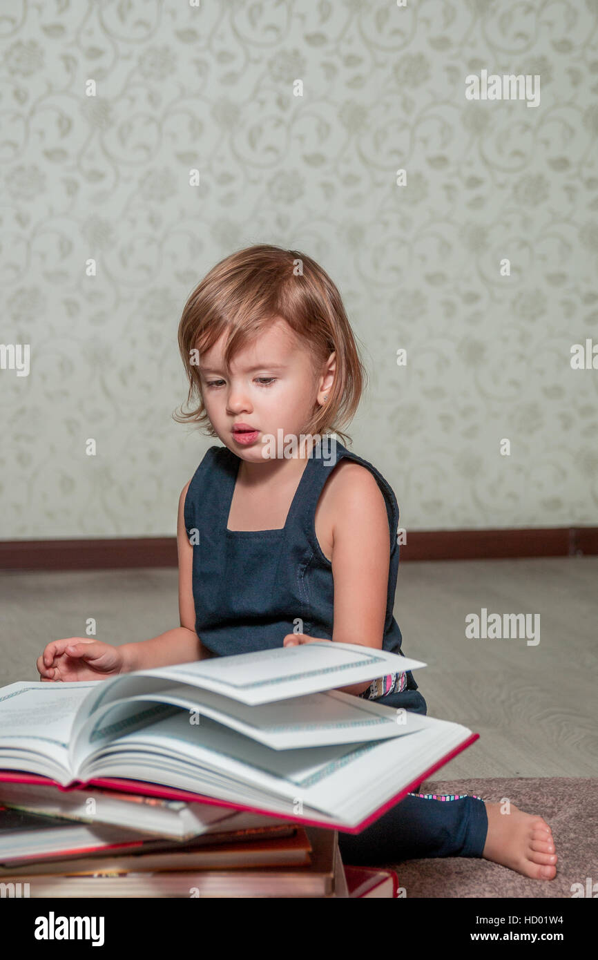 A little girl in a dark blue dress reading a book sitting on the floor ...