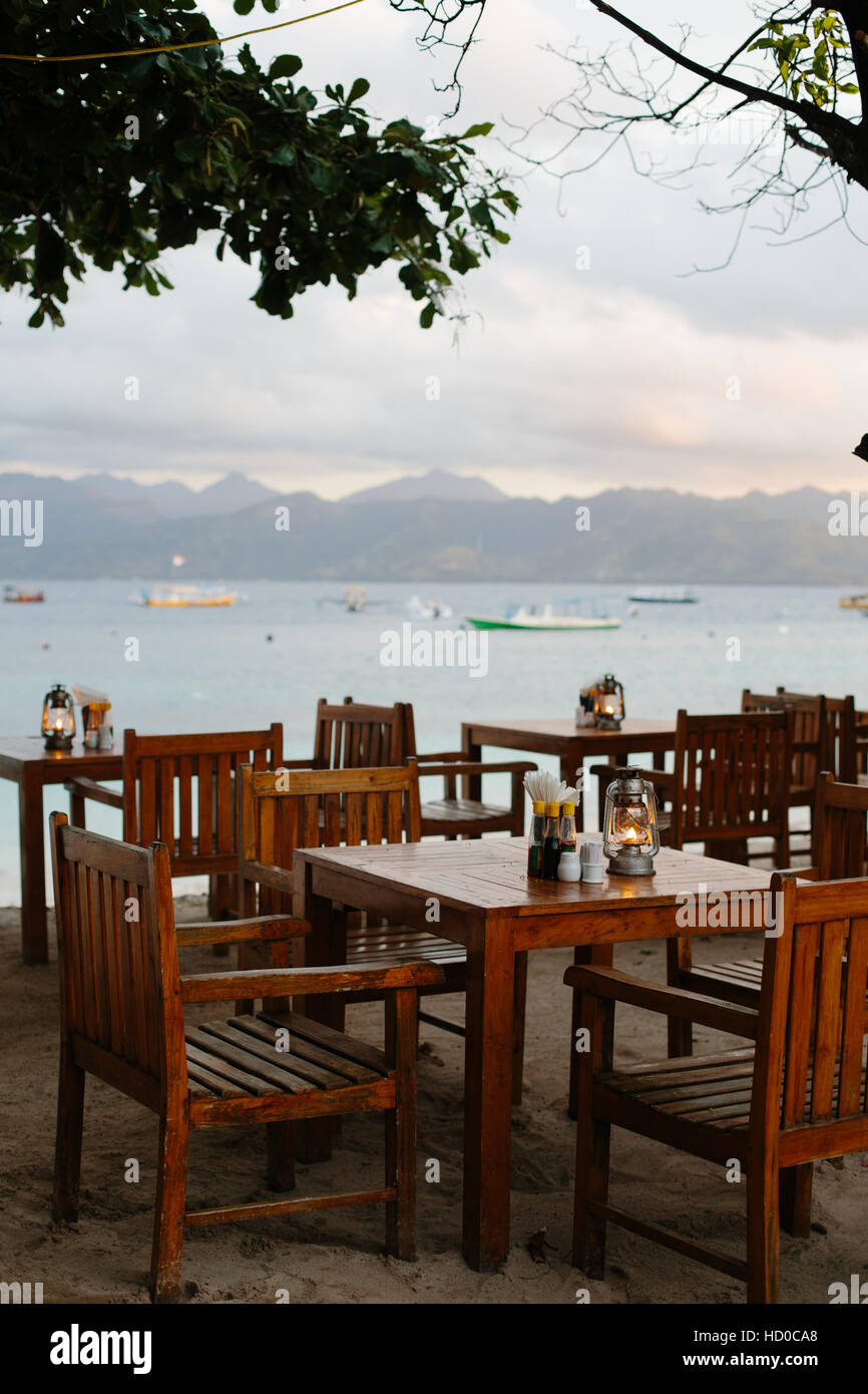 Empty wooden tables & chairs at a beachside restaurant on Gili Trawangan with a sunsetview of lombok and boats in the background Stock Photo