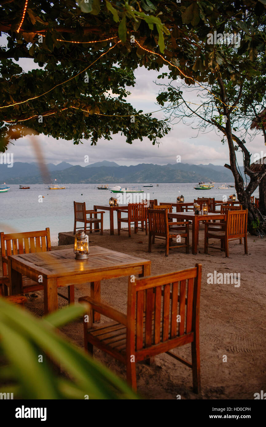 Wooden tables & chairs at a beachside restaurant in Gili Trawangan, with a view of Lombok and the beach and boats Stock Photo