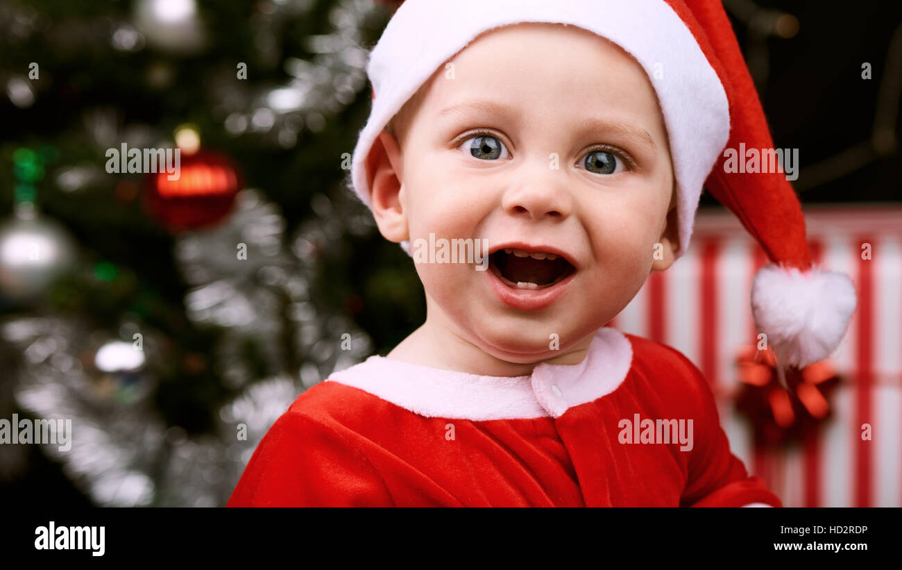 Happy cheerful expression on a baby boy's face while looking at the camera dressed in a red santa claus outfit and a christmas tree in the background. Stock Photo