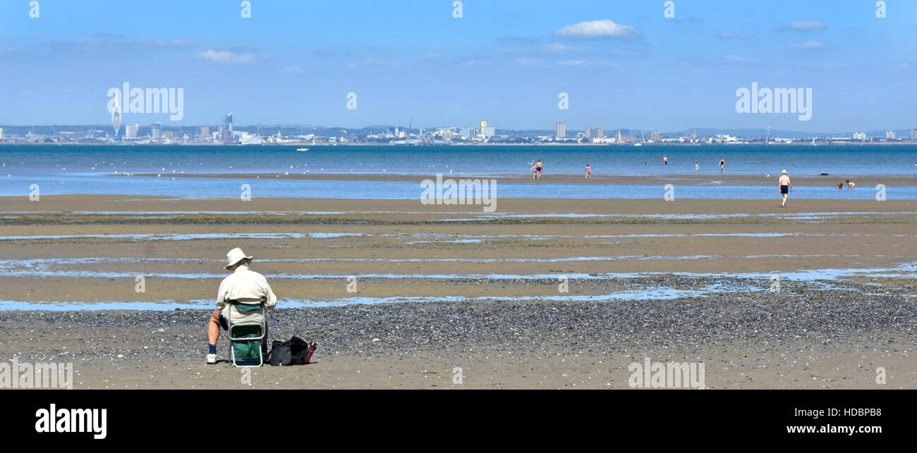 Back view of older man sitting alone on seaside beach chair sketching views of the Solent towards distant Portsmouth hot summer day Stock Photo