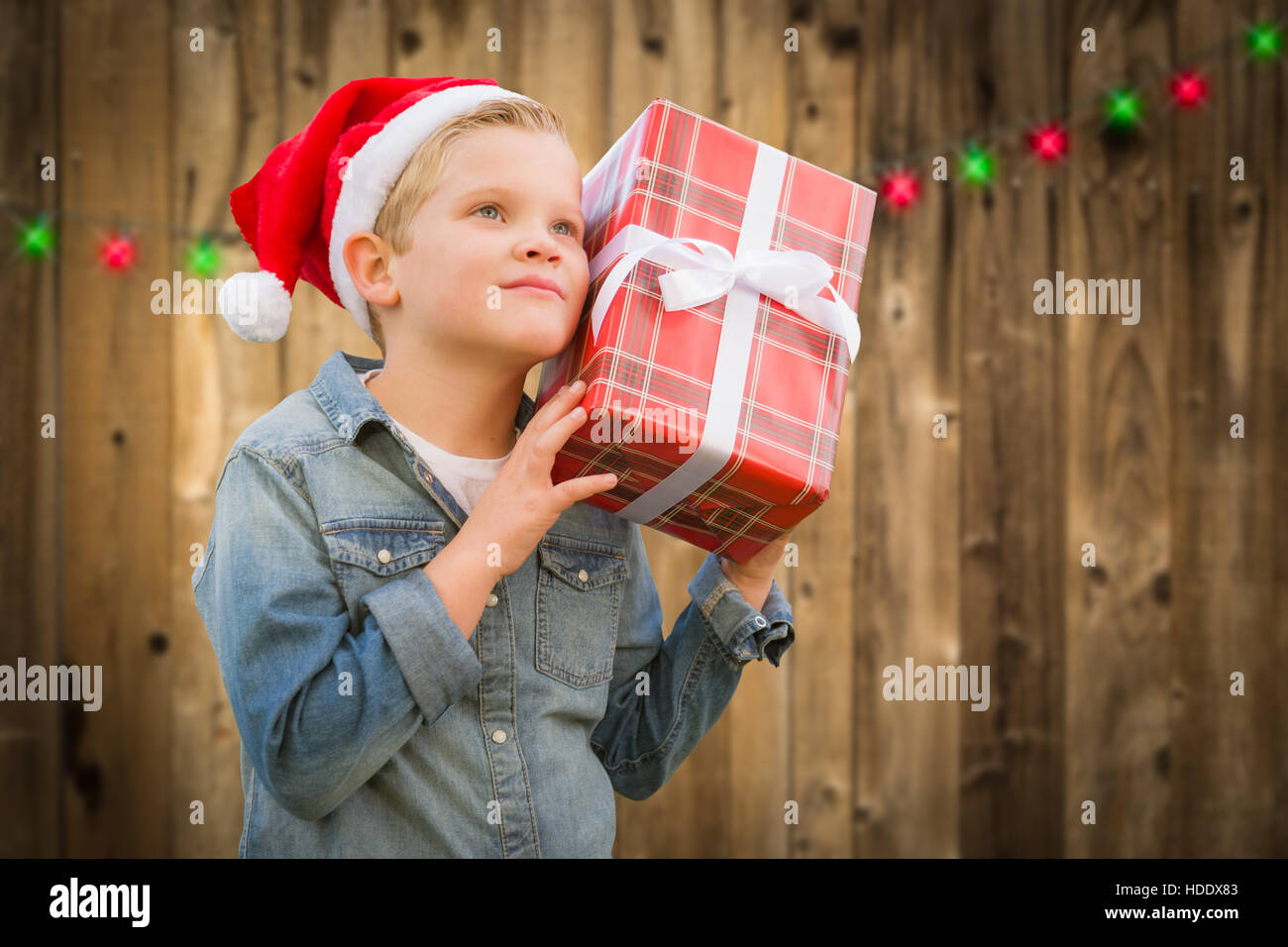 Curious Young Boy Wearing Santa Hat Holding Christmas Gift On A Wood Fence Background. Stock Photo