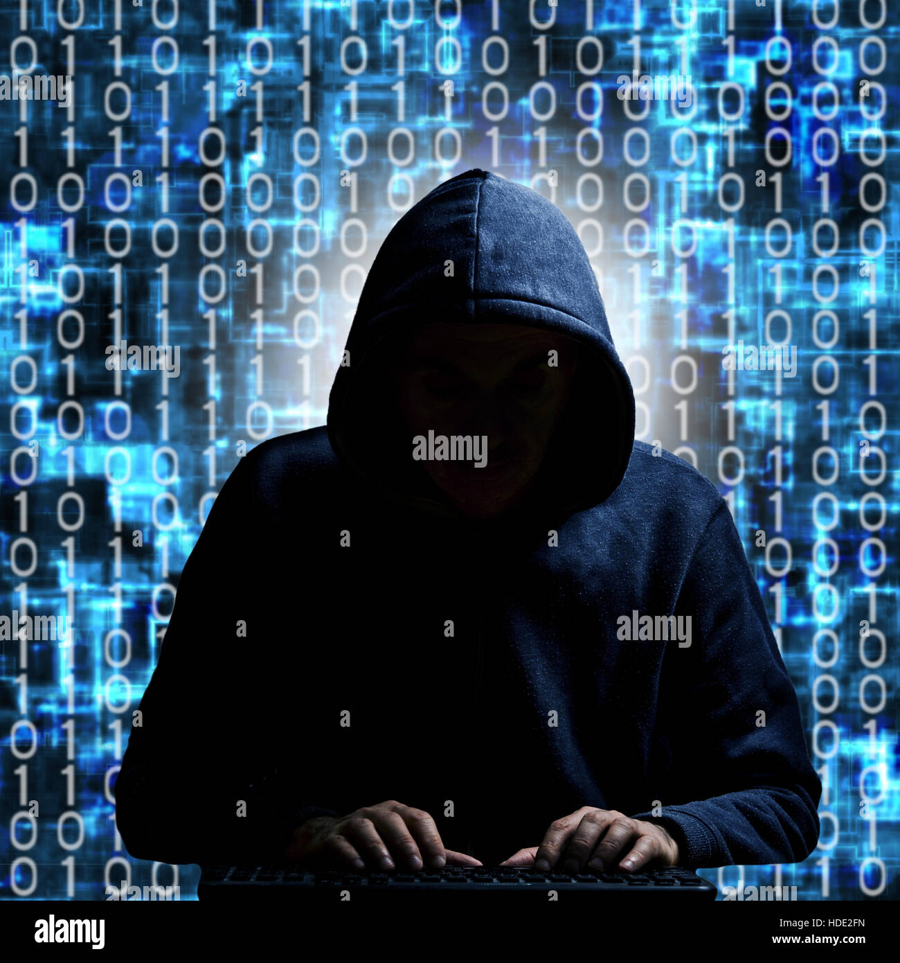 anonymous hacker in hood typing on a keyboard, with binary background Stock Photo