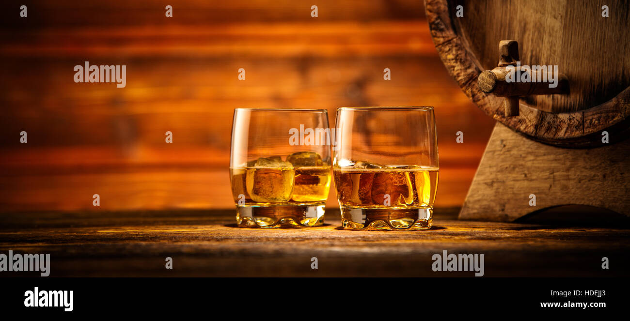 Two glasses of whiskey with ice cubes served on wooden planks with keg. Vintage countertop with highlight and a glass of hard liquor Stock Photo