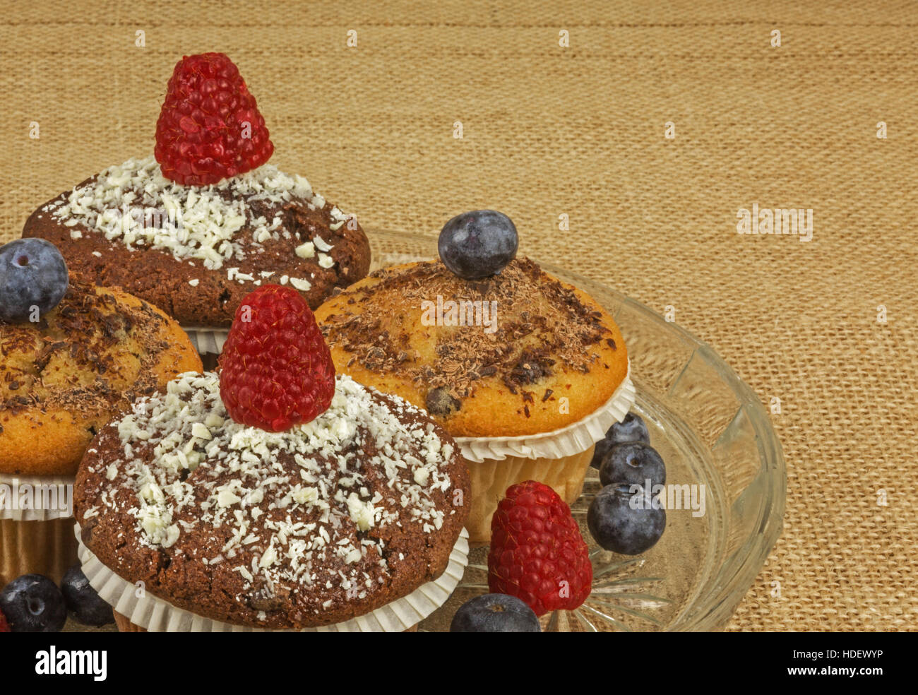 Four muffins sprinkled with grated chocolate and garnished with fresh raspberries and blueberries on a glass plate and the whole bob on the tablecloth Stock Photo