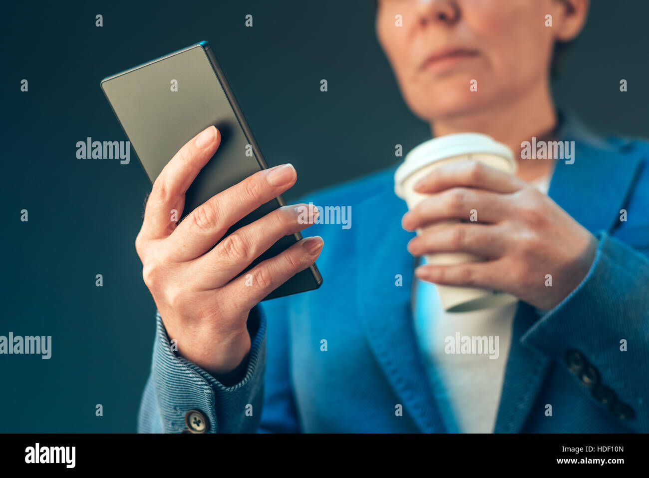 Satisfied young adult businesswoman drinking coffee to go and looking at the mobile phone Stock Photo