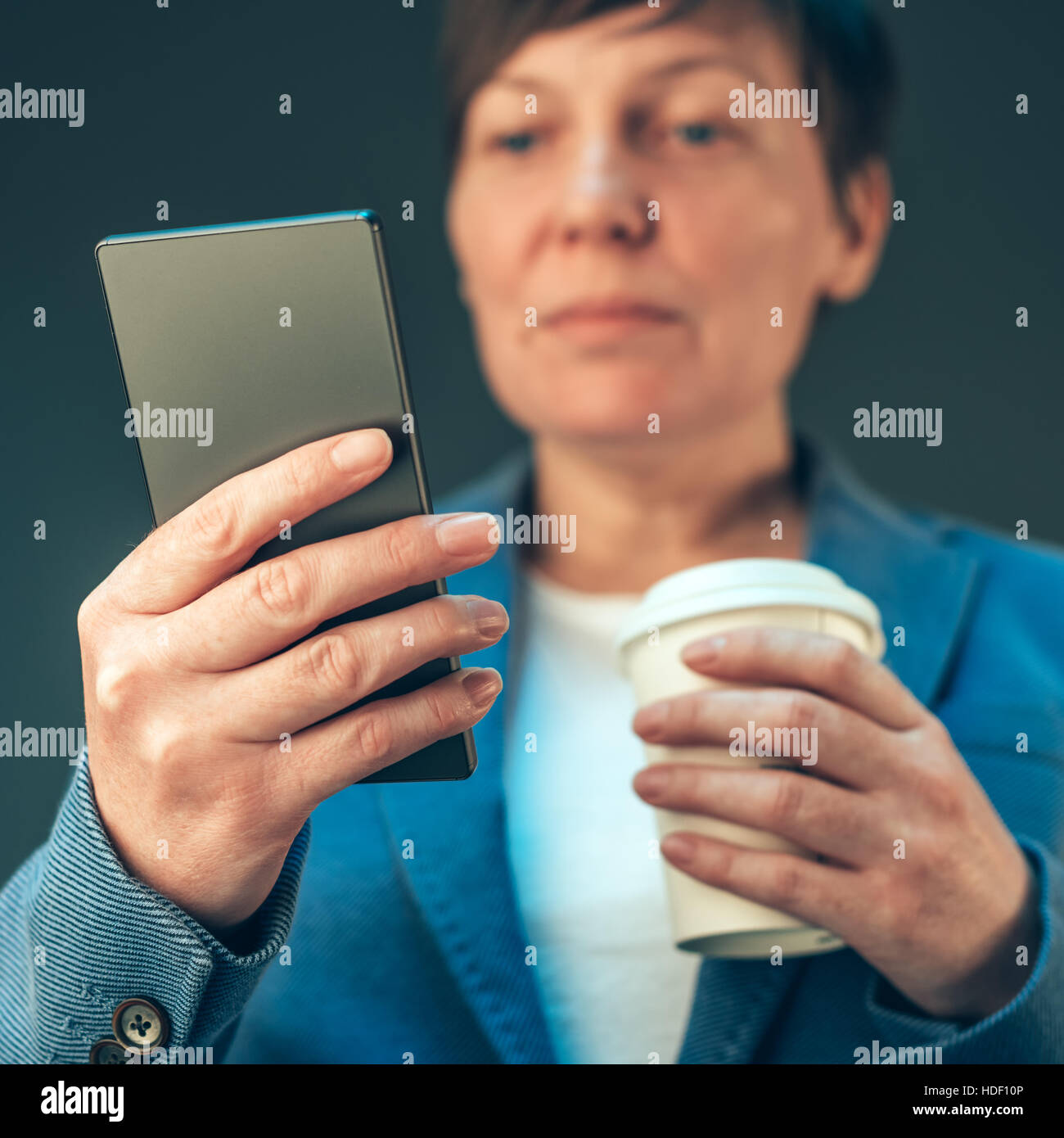 Satisfied young adult businesswoman drinking coffee to go and looking at the mobile phone Stock Photo