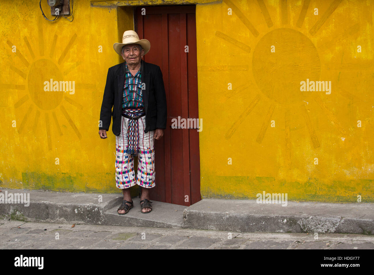 An older Mayan man in traditional dress of San Pedro la Laguna, Guatemala, stands in front of brightly painted house with a red door and yellow sun de Stock Photo