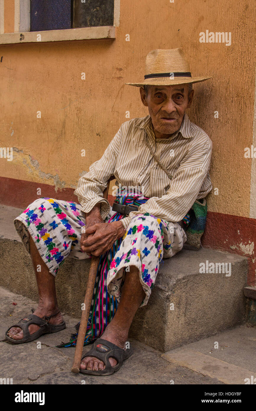 93 year old Mayan man in traditional dress sits on street in San Pedro la Laguna, Guatemala. Sitting on the curb with his cane in front of a pastel pa Stock Photo