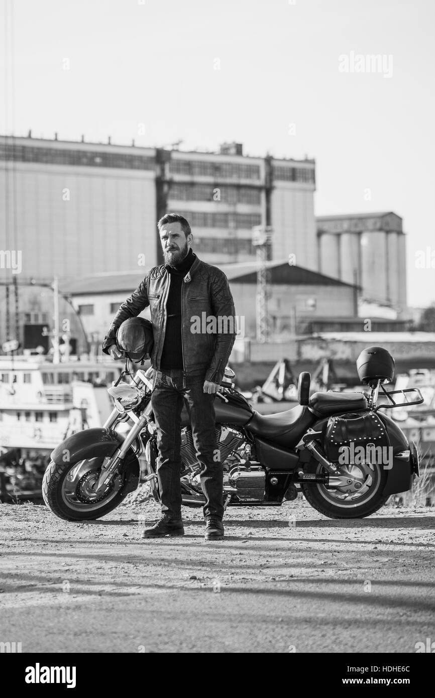 Full length portrait of biker holding helmet while standing by motorcycle against industrial setting Stock Photo