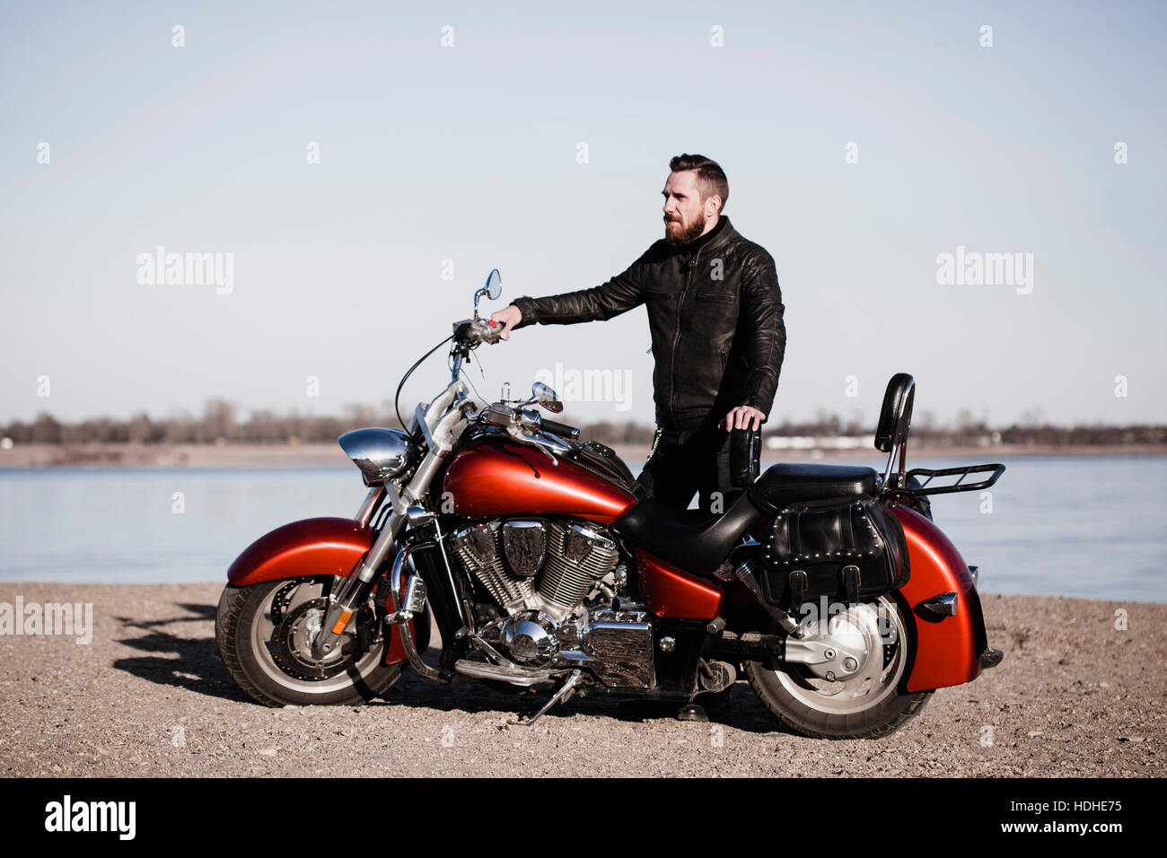 Full length portrait of biker standing by motorcycle and looking away at lakeshore Stock Photo