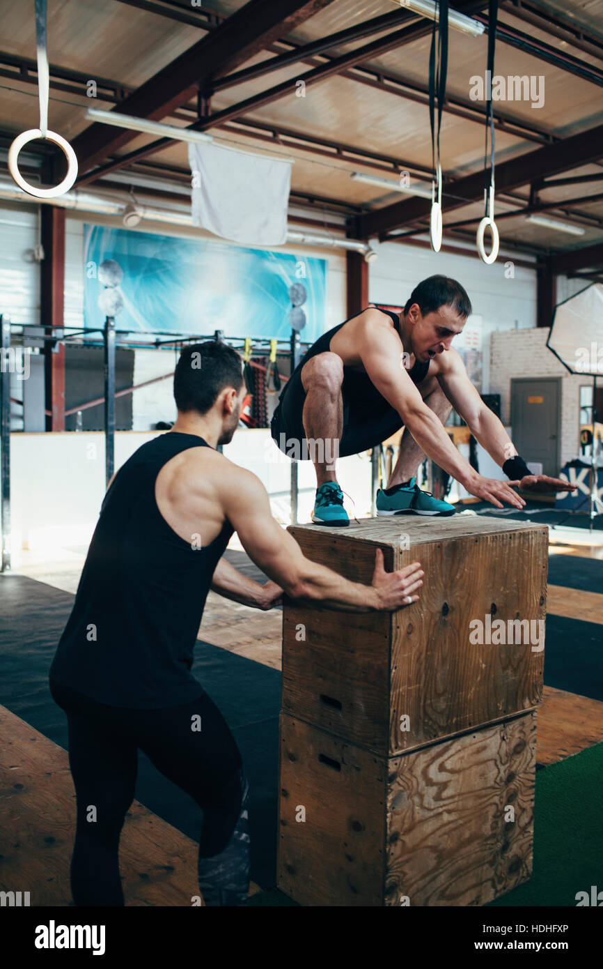 Male athlete assisting friend in doing box jumping at gym Stock Photo