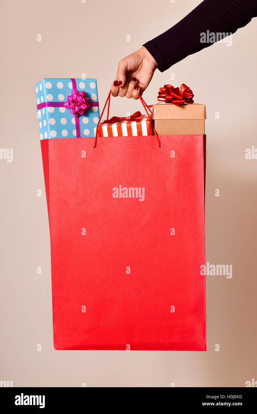 closeup of the hand of a young caucasian woman with her fingernails painted red holding a red shopping bag full of gifts wrapped in different papers, Stock Photo