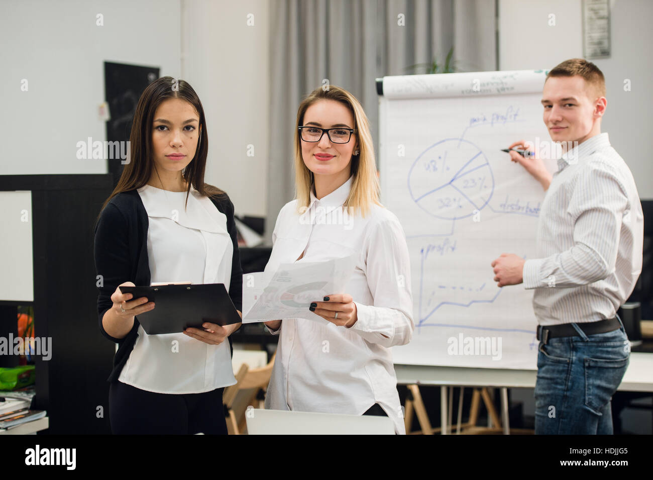 Two beautiful office workers having conversation while man colleague drawing business strategy on flip chart. Stock Photo