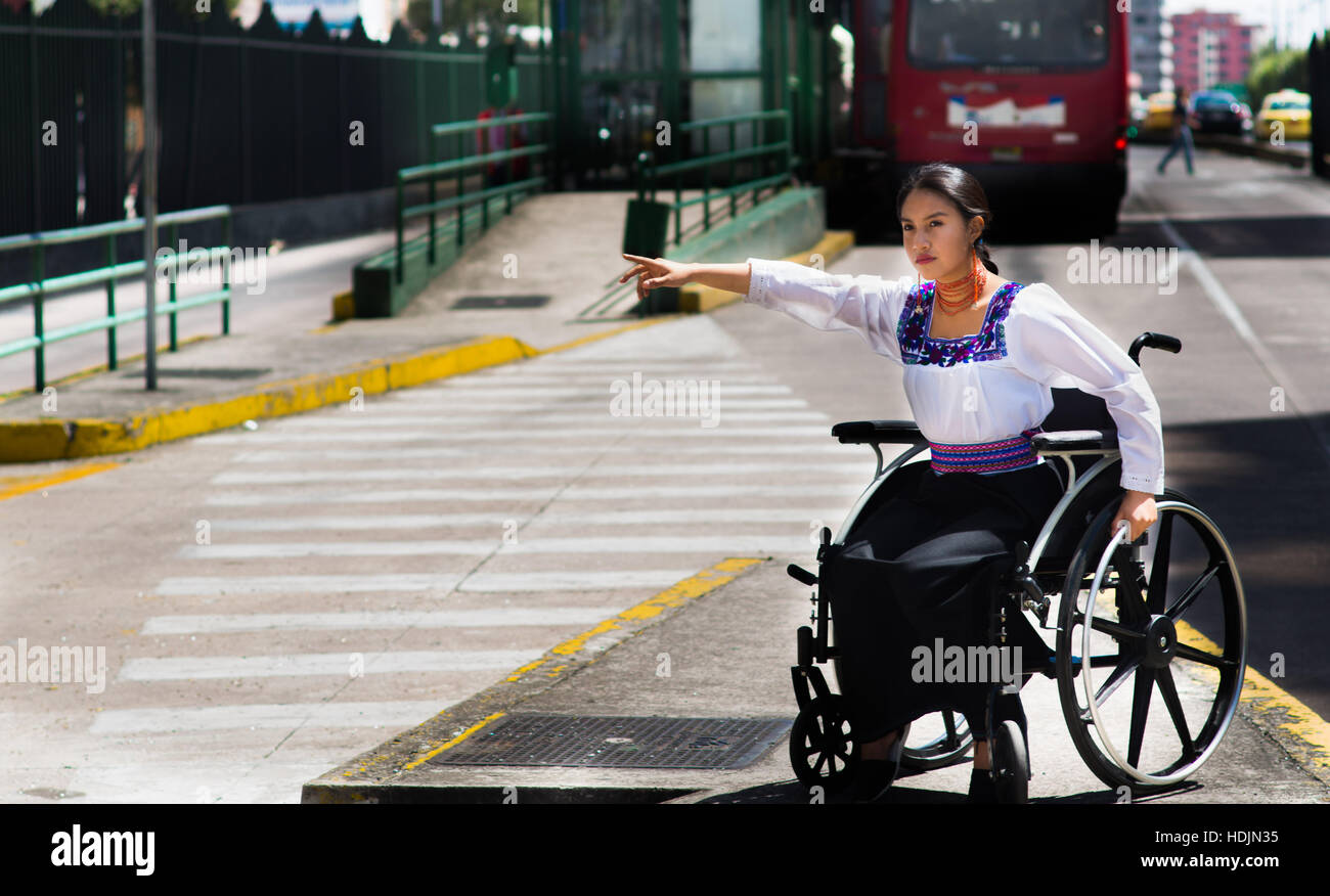 Young brunette woman sitting in wheelchair smiling with positive attitude, holding out arm looking for taxi, outdoors environment, physical recovery concept Stock Photo