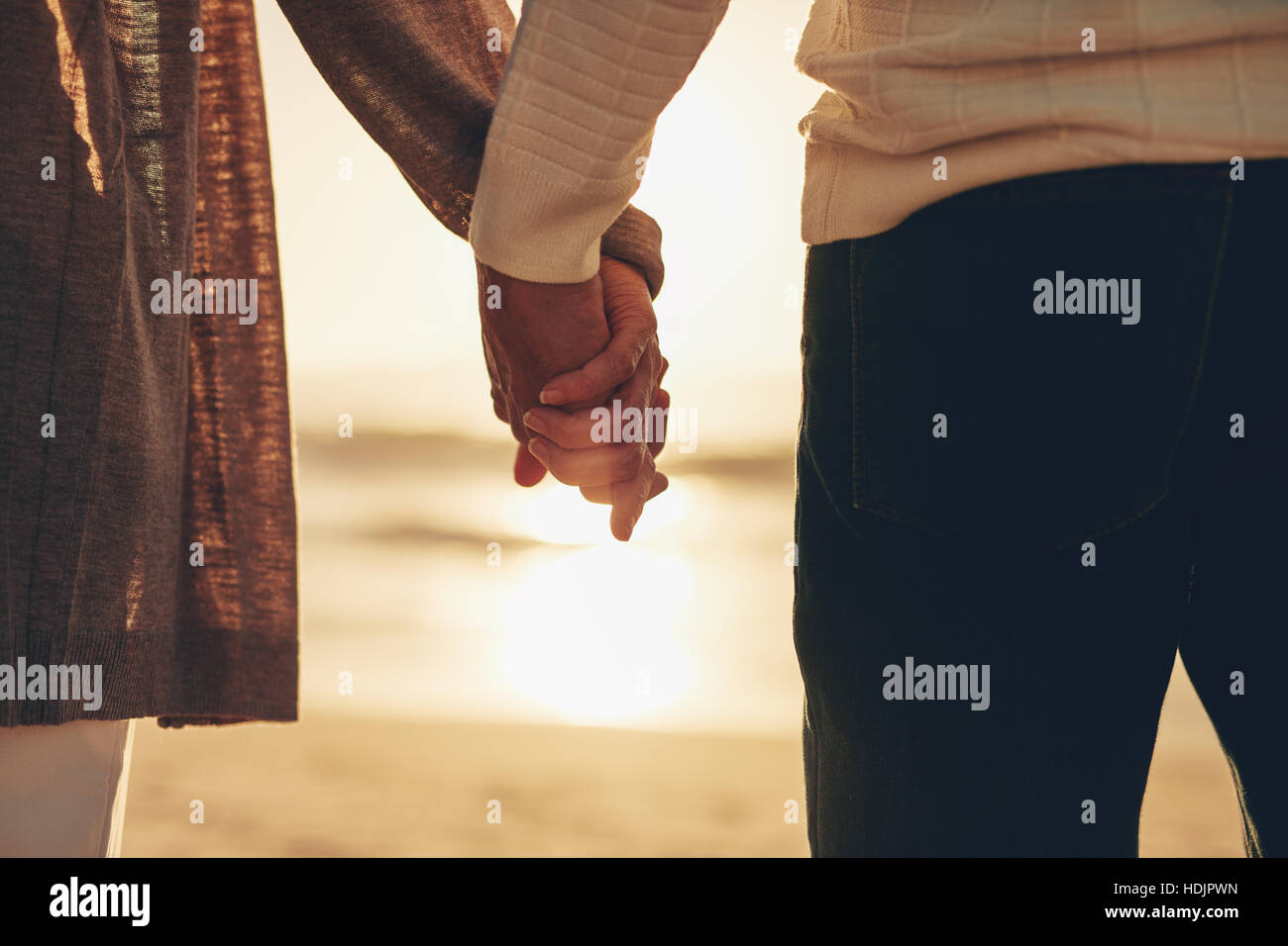 Rear view close up shot of a senior couple holding hands at the ocean during sunset. Stock Photo