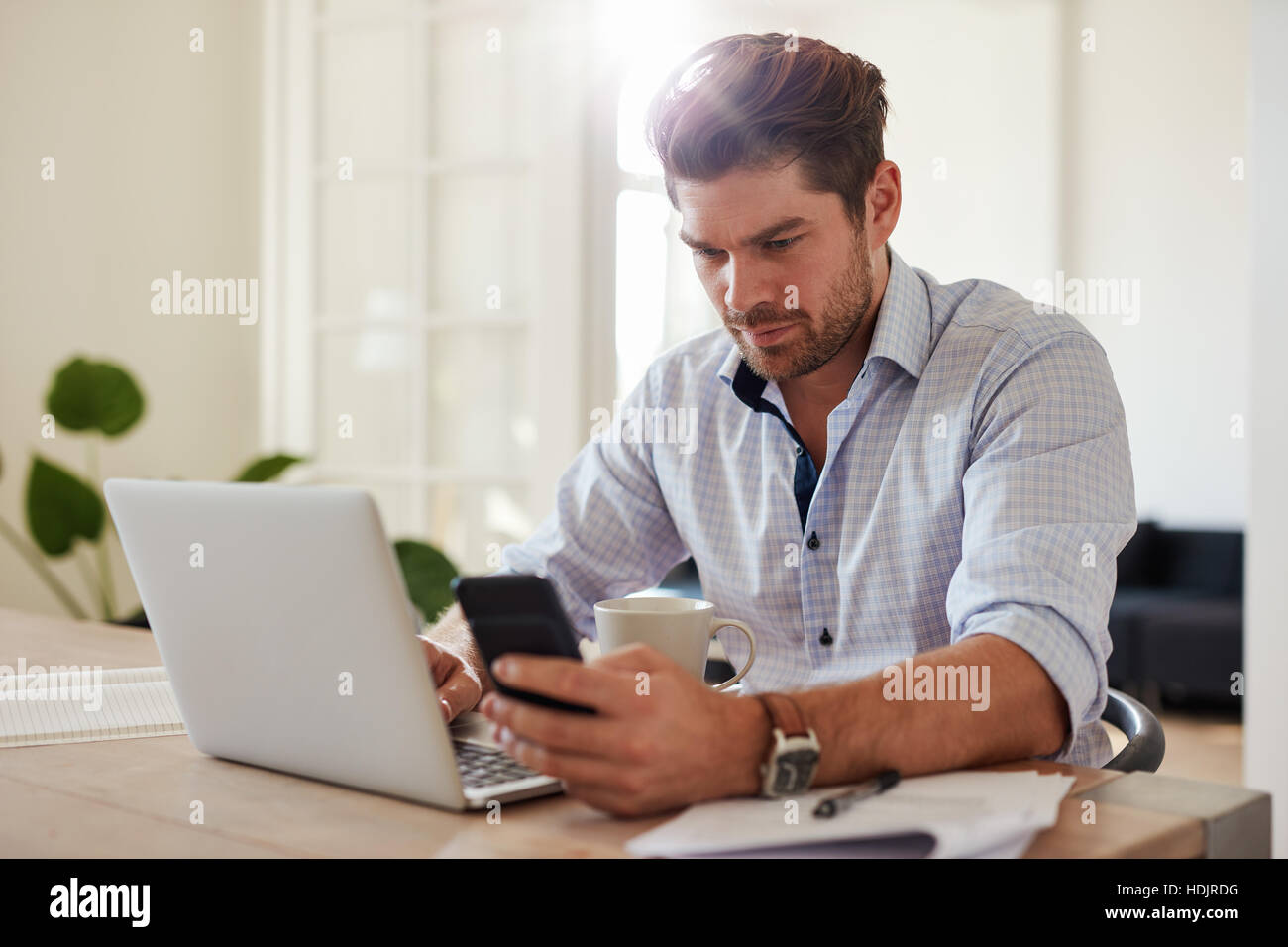 Shot of young man working with laptop and mobile phone at his home office. Handsome male sitting at table holding smartphone and using laptop computer Stock Photo