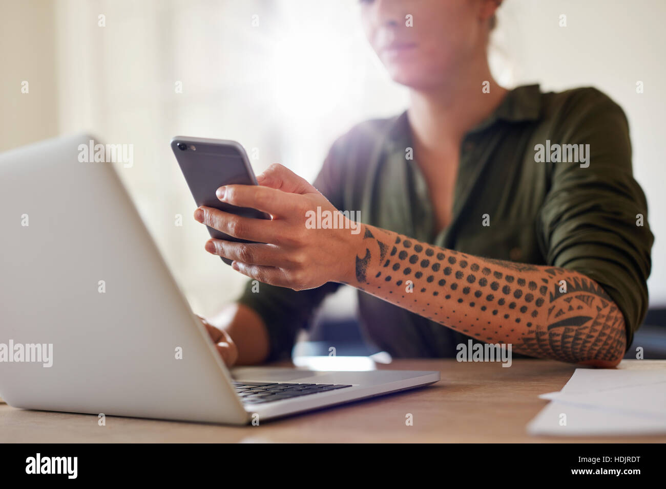 Shot of young woman using mobile phone while working on laptop. Focus on smart phone in hand of a woman. Stock Photo