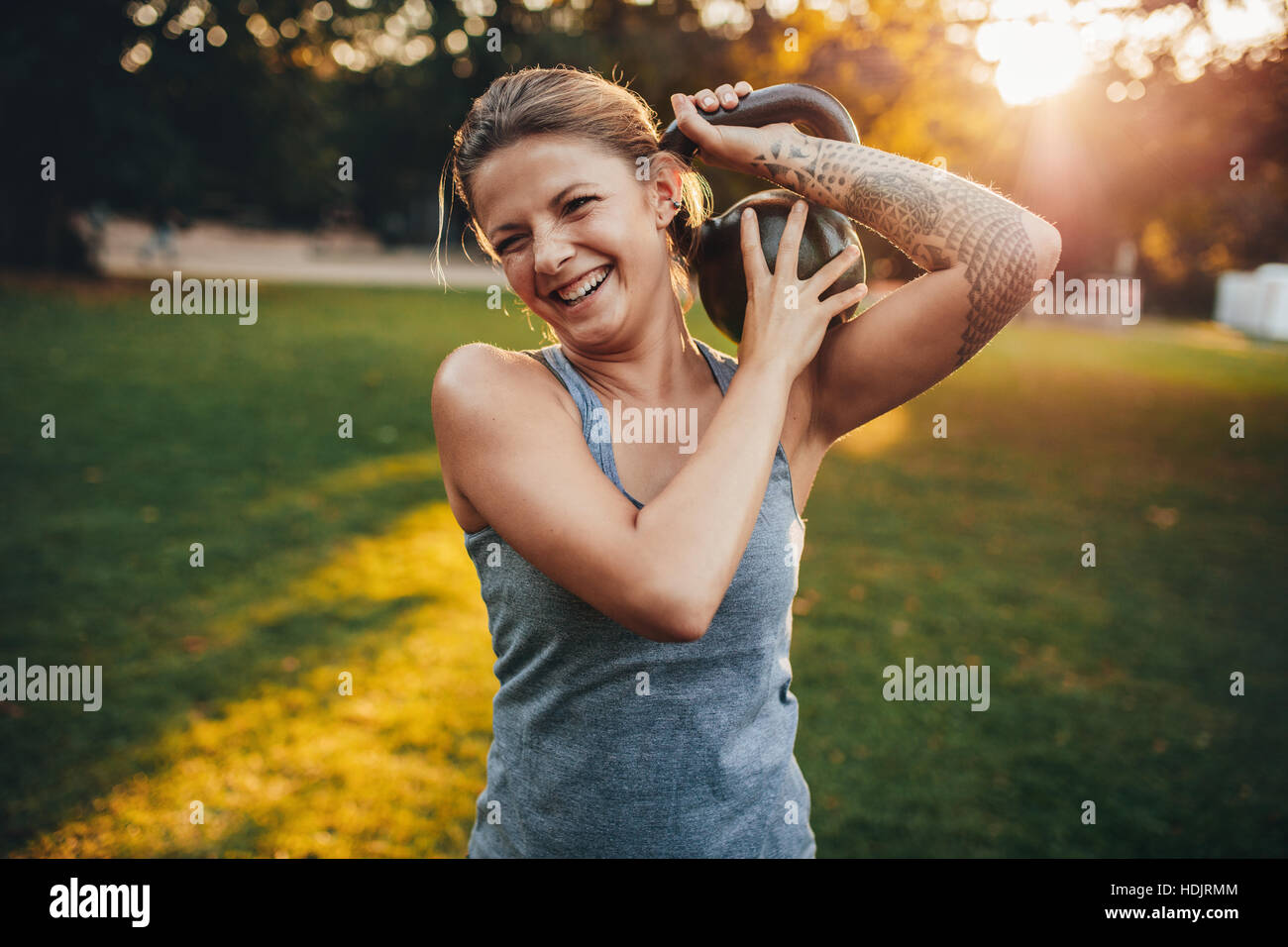 Portrait of happy young woman with kettlebell weights on her shoulder in the park. Stock Photo