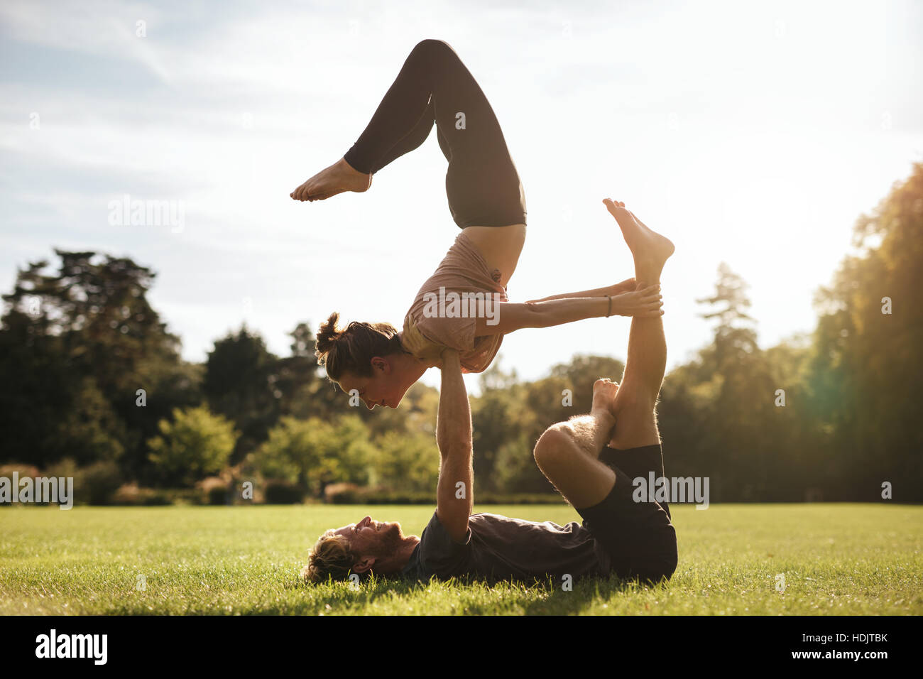 Fit young couple doing acroyoga on grass. Man and woman in park practising pair yoga poses in sunny morning. Stock Photo